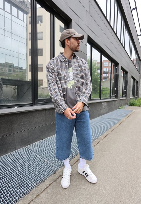 Mann mit grauem Cap, kariertem Hemd, bedrucktem T-Shirt, kurzen blauen Jeans, weißen Socken und weißen Turnschuhen, stehend auf einem Bürgersteig in der Stadt in der Nähe von Glasfenstern.