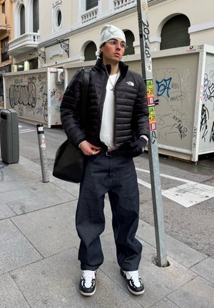 Joven con chaqueta acolchada negra, gorro blanco, gafas, jeans oscuros holgados y zapatillas negras y blancas, de pie en una acera urbana con grafiti.