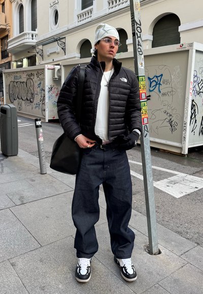 Joven con chaqueta acolchada negra, gorro blanco, gafas, jeans oscuros holgados y zapatillas negras y blancas, de pie en una acera urbana con grafiti.