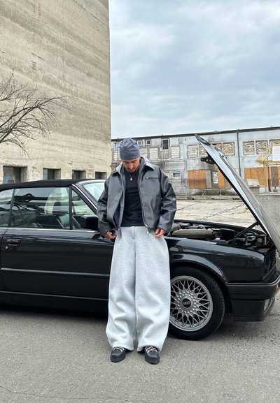 Chaqueta bomber de cuero negra, pantalones de chándal gris oversize, calzado negro. De pie junto a un coche negro con el capó abierto y llantas detalladas.