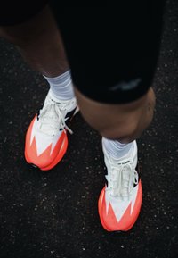 Jambes portant un short noir, des chaussettes blanches et des chaussures de course blanc éclatant et orange néon sur une surface asphaltée.