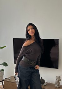 Haut marron à manches longues et épaules dénudées, associé à un pantalon large en jean foncé, complété par une ceinture noire épaisse et un sac texturé.