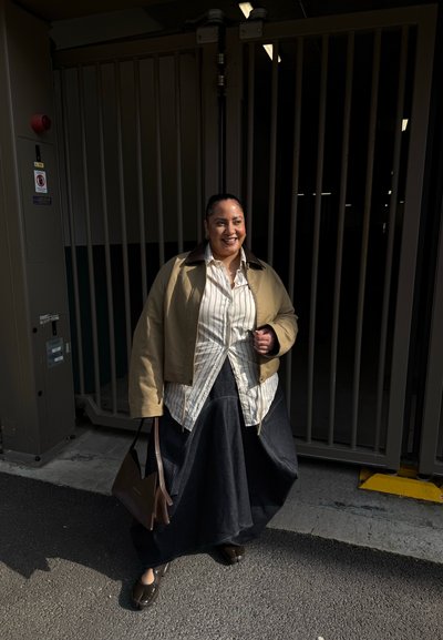 Mujer sonriente de pie frente a una entrada con reja, viste chaqueta beige, camisa a rayas, falda larga negra y sostiene un bolso marrón.
