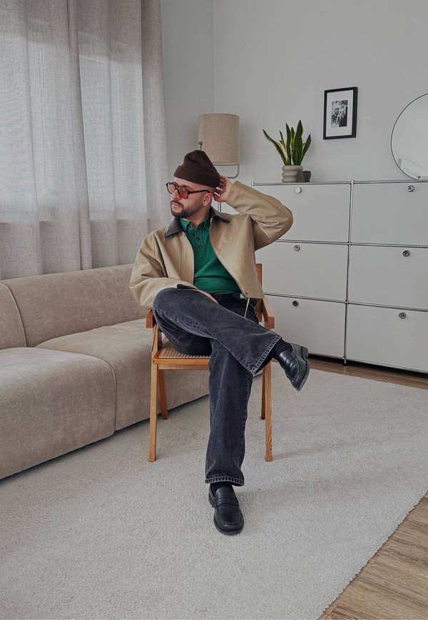 Mann mit beiger Jacke, grünem Hemd, schwarzer Mütze und Brille sitzt mit verschränkten Beinen auf einem Holzstuhl in einem modernen Wohnzimmer.