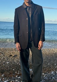 Blazer noir à motifs avec fermeture éclair en cuir sur le devant, assorti à un jean ample et foncé. Le blazer possède un col et des boutons sur le devant.