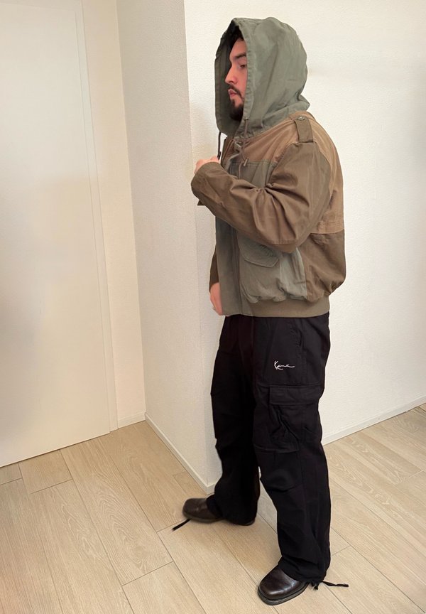 Uomo con indosso una giacca marrone bicolore con cappuccio alzato, pantaloni cargo neri e scarpe nere, in piedi all'interno vicino a una parete bianca e a una porta.