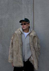 Faux bontjas in licht beige met een zachte, ruige textuur; gedragen over een grijze hoodie met ritssluiting. Zwarte jeans en een groene pet maken de look af.