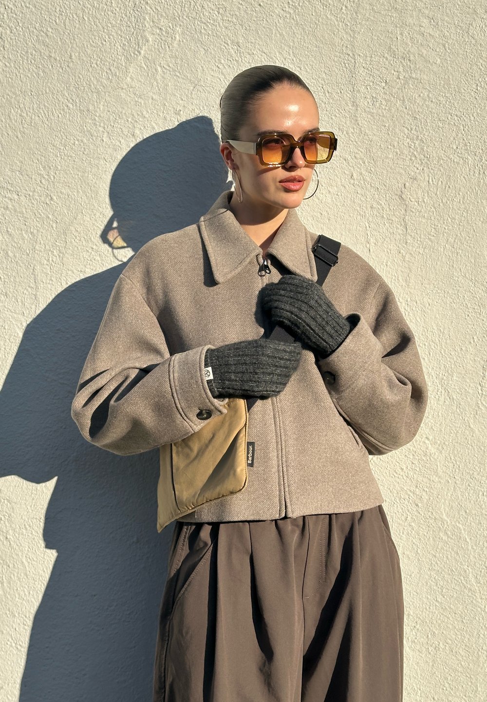 Veste en laine marron avec col, fermeture éclair, associée à des gants gris côtelés et un sac bandoulière beige. Le mannequin pose devant un mur clair.
