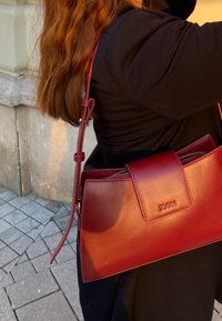 Κόκκινη δερμάτινη τσάντα χειρός με δομημένο σχέδιο, κλείσιμο με φερμουάρ και ρυθμιζόμενο λουρί ώμου. Διαθέτει ανάγλυφο λογότυπο στην μπροστινή πλευρά.