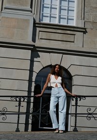 White sleeveless top featuring a deep v-neck, light blue high-waisted jeans, and pale heels. Accompanied by a light-coloured handbag and wavy hair, set against a grey stone wall.