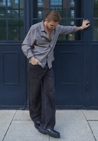 Man in een grijs shirt en zwarte wijde broek leunt tegen een donkerblauwe deur, kijkt omlaag met één hand in zijn zak en de andere hand op de deur.