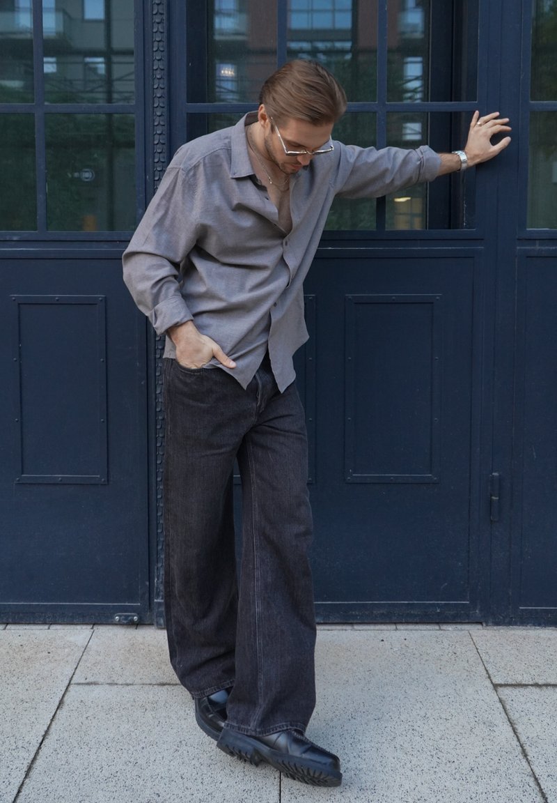 Uomo vestito con una camicia grigia e pantaloni larghi neri si appoggia a una porta blu scuro, guardando in basso con una mano in tasca e l'altra sulla porta.