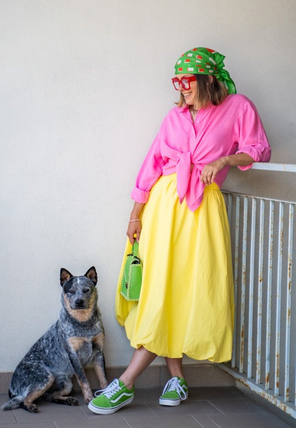 Frau in farbenfroher Kleidung mit rosa Hemd, gelbem Rock, grünem Kopftuch und Schuhen lehnt am Geländer neben einem sitzenden Blue Heeler Hund.