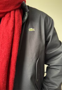 Črna vodoodporna jakna z zelenim logoom krokodila, v kombinaciji z debelim rdečim šalom iz teksturirane tkanine. Ima žep s gumbu.