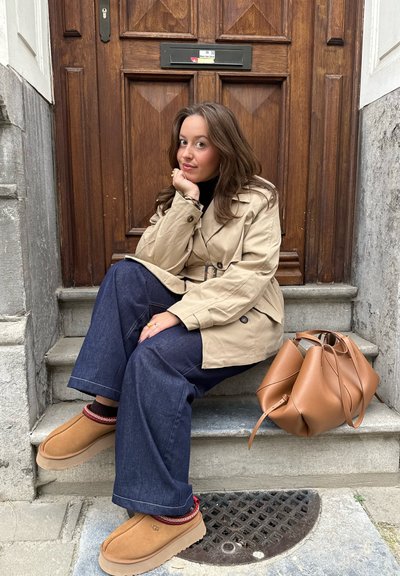 Gabardina beige con cintura ceñida, vaqueros de pierna ancha azul oscuro, zapatos de ante marrón con un acento rojo, y un bolso de cuero claro en los escalones.