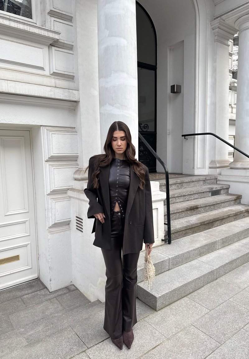 Femeie îmbrăcată într-un top și pantaloni negri din piele, cu un sacou negru, ținând o geantă bej împletită, stând lângă scările de la intrarea într-o clădire cu coloane albe.