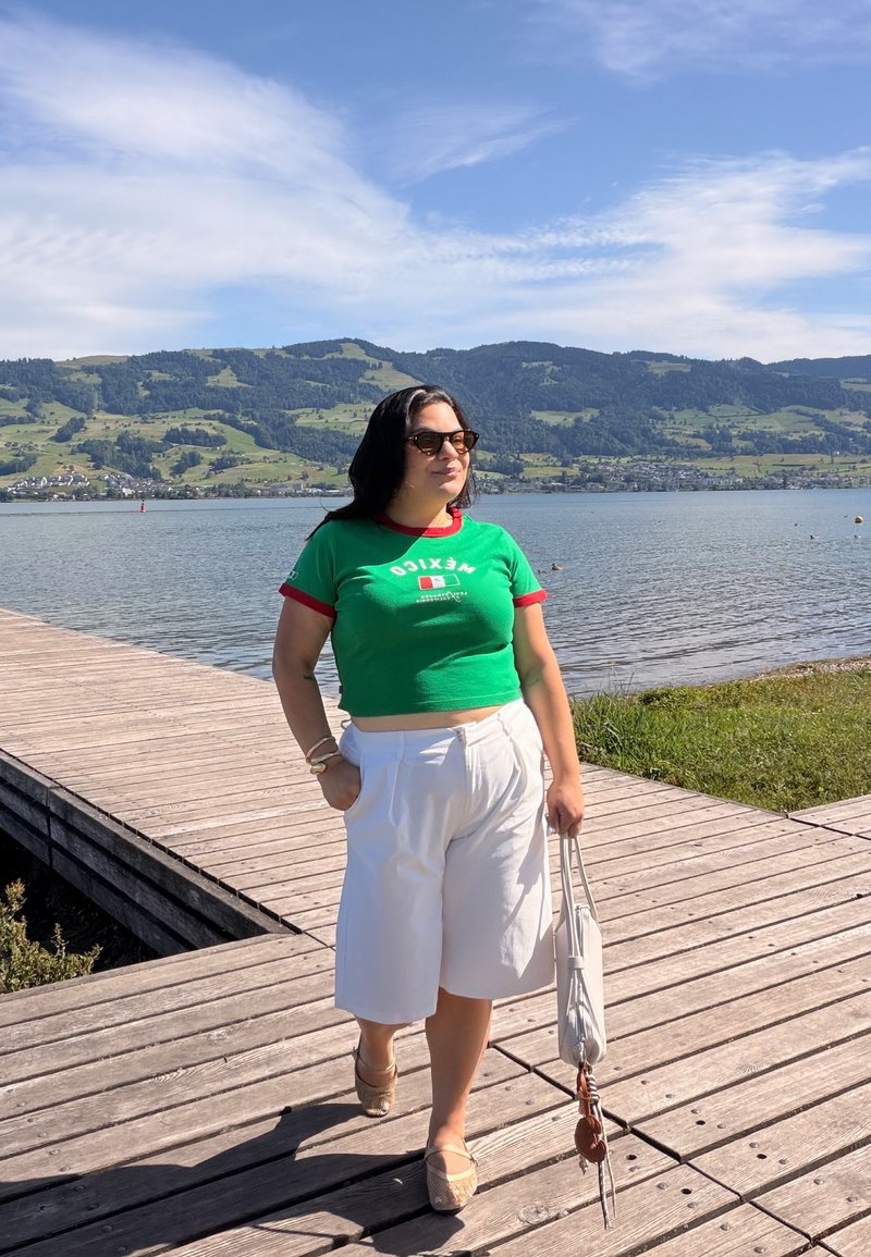 Femme portant un t-shirt vert du Mexique et un short blanc, debout sur une jetée en bois près d'un lac avec des collines et un ciel bleu en arrière-plan.