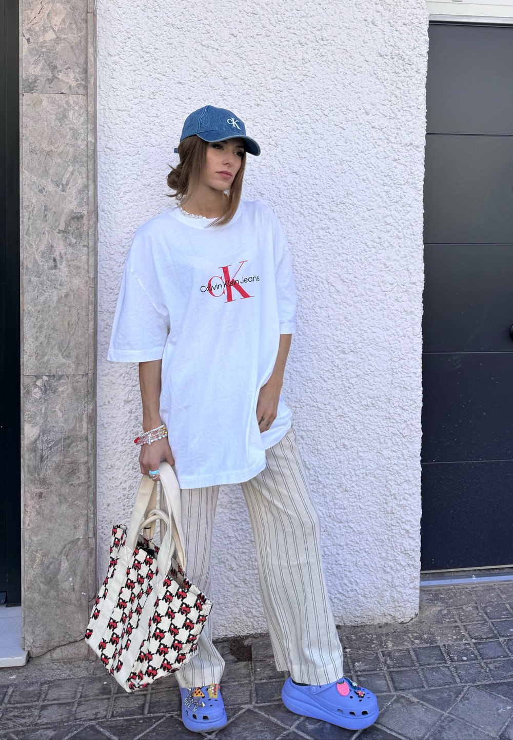 Overdimensioneret hvid t-shirt med rødt 'Calvin Klein Jeans' logo, stribede beige bukser, blå træsko med charms og en mønstret tote bag.