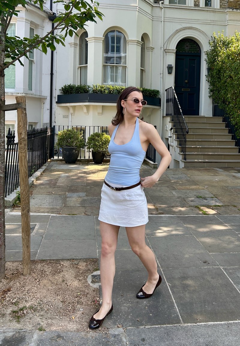 Femme portant des lunettes de soleil, un haut bleu clair, une jupe blanche et des chaussures noires marchant sur un trottoir pavé près d’un immeuble résidentiel avec des escaliers et des plantes.