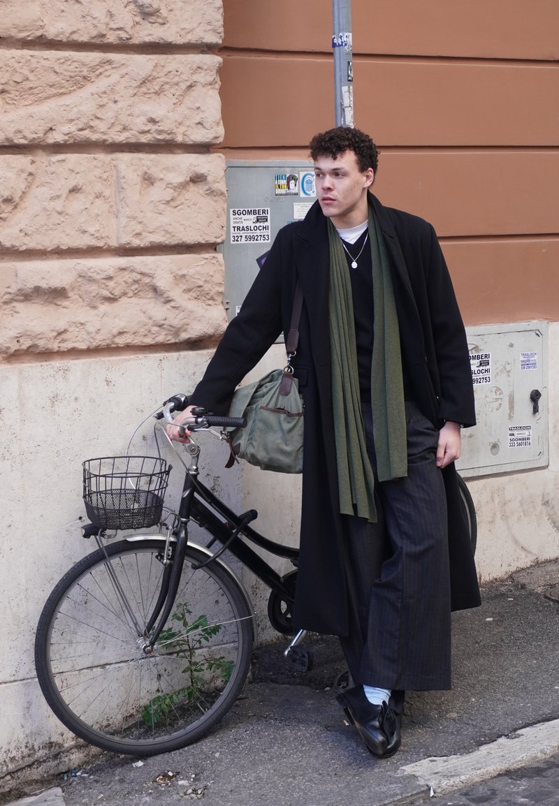 Jeune homme en long manteau noir et écharpe verte appuyé sur un vélo noir avec panier à un coin de rue urbain, devant des murs en pierre et stuc.