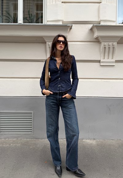 Camisa de botón de denim oscuro con mangas largas, combinada con jeans de pierna ancha y botas negras de punta afilada. Se incluye un bolso de hombro.