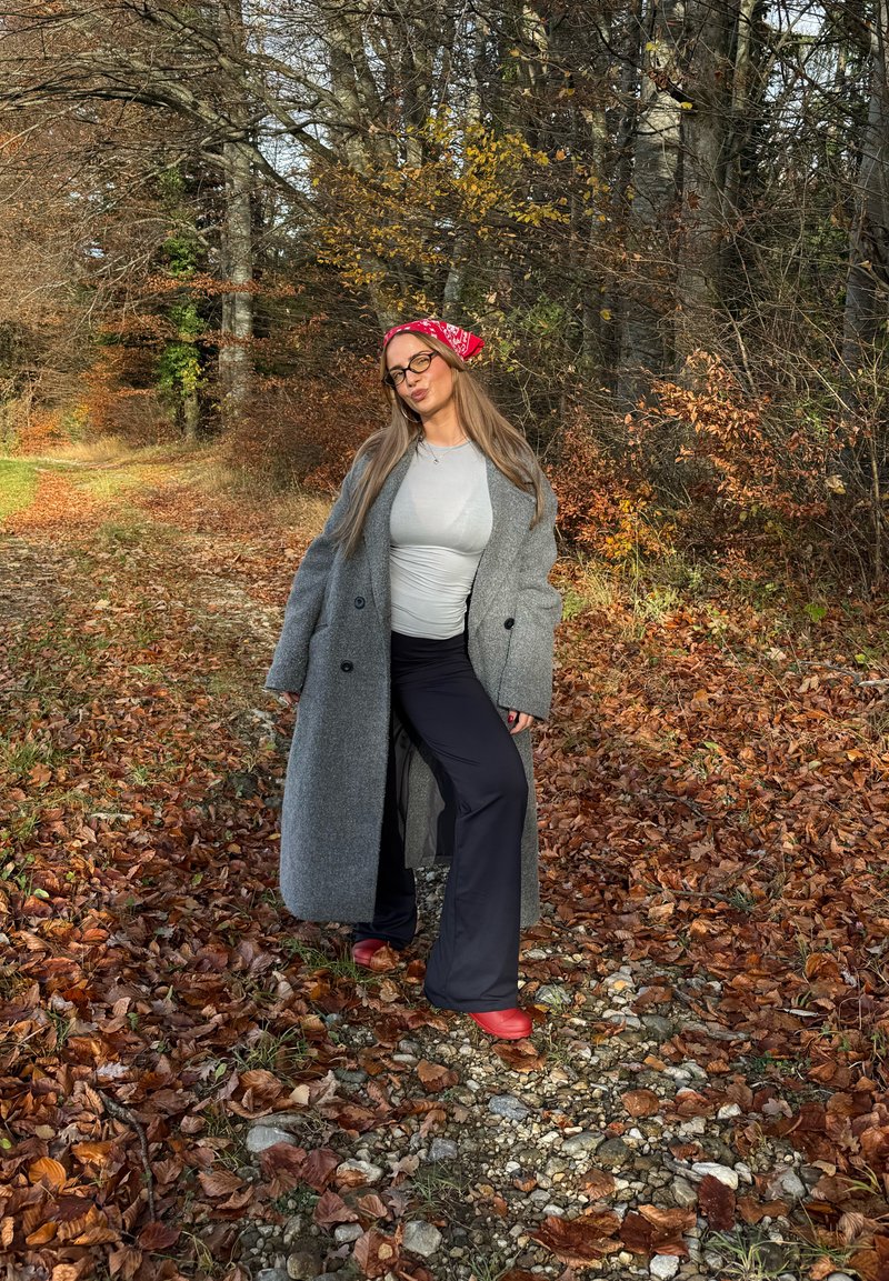 Long gray wool coat, gray shirt, black pants, and red shoes. Red patterned scarf tied around the hair. Autumn leaves and trees in background.