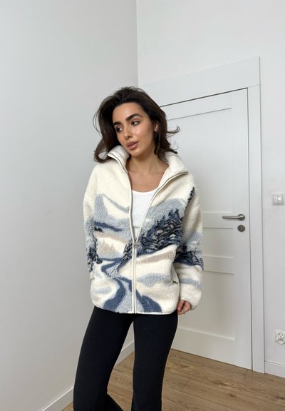 Chaqueta de forro polar en crema con estampado de paisaje montañoso azul, cierre completo con cremallera, cuello alto y bolsillos laterales, combinada con una camisa blanca y pantalones negros.