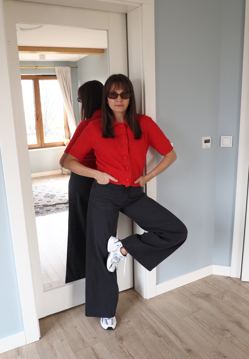 Vrouw met zonnebril, rode korte mouwen blouse met knopen, zwarte broek met wijde pijpen en witte sneakers, poserend binnen met één opgeheven been.