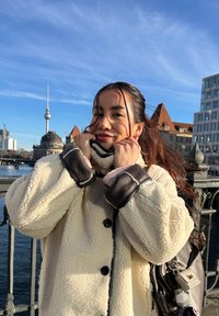 Young woman in cream sherpa coat adjusting collar on bridge with river and city buildings, including the Berlin TV Tower, under a blue sky.