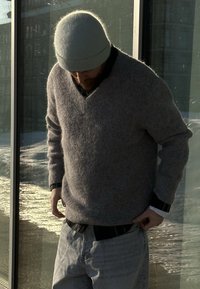 Homme portant un pull en maille gris et un bonnet ajustant son jean délavé près d'une fenêtre en verre éclairée par le soleil.