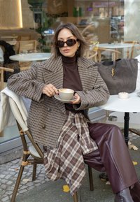 Brązowy płaszcz dwurzędowy w jodełkę, brązowy golf, spódnica w kratę i bordowe skórzane spodnie. Czarne duże okulary przeciwsłoneczne, trzymająca filiżankę.