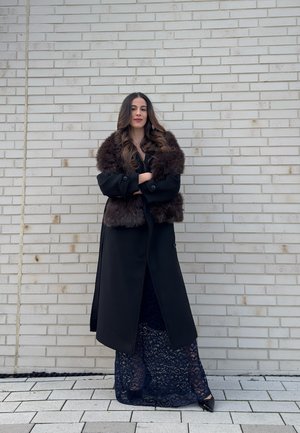 Mujer con cabello largo y ondulado de pie contra una pared de ladrillo claro, vistiendo un abrigo negro con un gran cuello de piel y una falda de encaje azul marino, con los brazos cruzados.