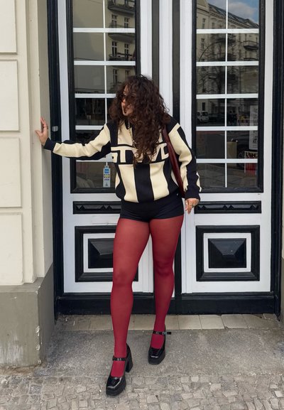 Suéter de rayas blancas y negras, medias rojas, zapatos de plataforma negros y un bolso marrón, de pie cerca de puertas dobles con paneles de vidrio.