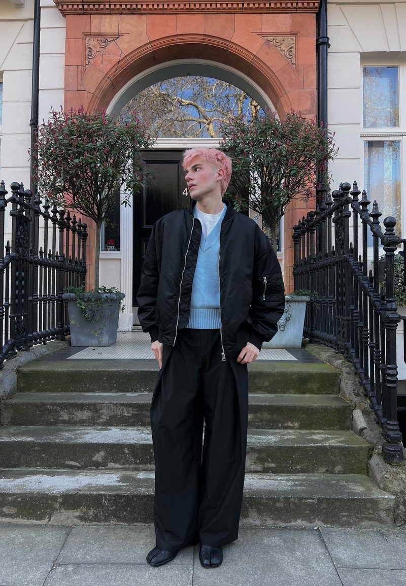 Nuori henkilö, jolla on vaaleanpunaiset hiukset, musta takki ja leveät mustat housut, seisoo kivisillä portailla rakennuksen ulkopuolella, jossa on kaareva ovi ja ruukkuistutuksia puita.