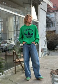 Πράσινο πλεκτό πουλόβερ με λογότυπο Adidas σε ναυτικό χρώμα, χαλαρής εφαρμογής μπλε τζιν παντελόνι και πράσινα παπούτσια slip-on, σε εξωτερικό αστικό περιβάλλον.