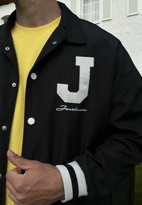 Černá bunda s bílou filcovou nášivkou "J" a vyšitým nápisem "Jacobsen" na hrudi, nošená přes žlutou košili. Rukávy s černobílými pruhy.