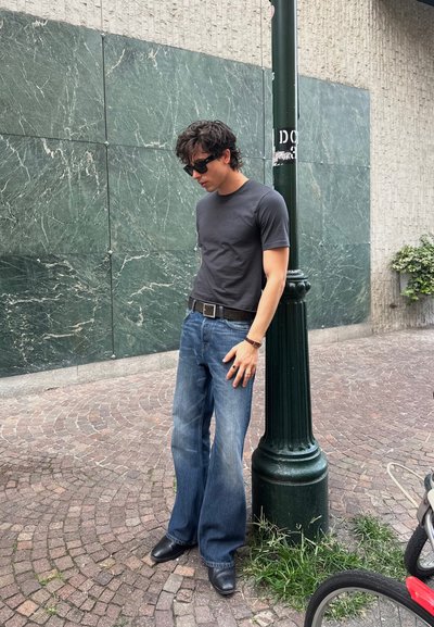 Joven con gafas de sol negras, camiseta gris oscuro, jeans azules holgados y zapatos negros apoyado en un farol verde en una calle de adoquines.