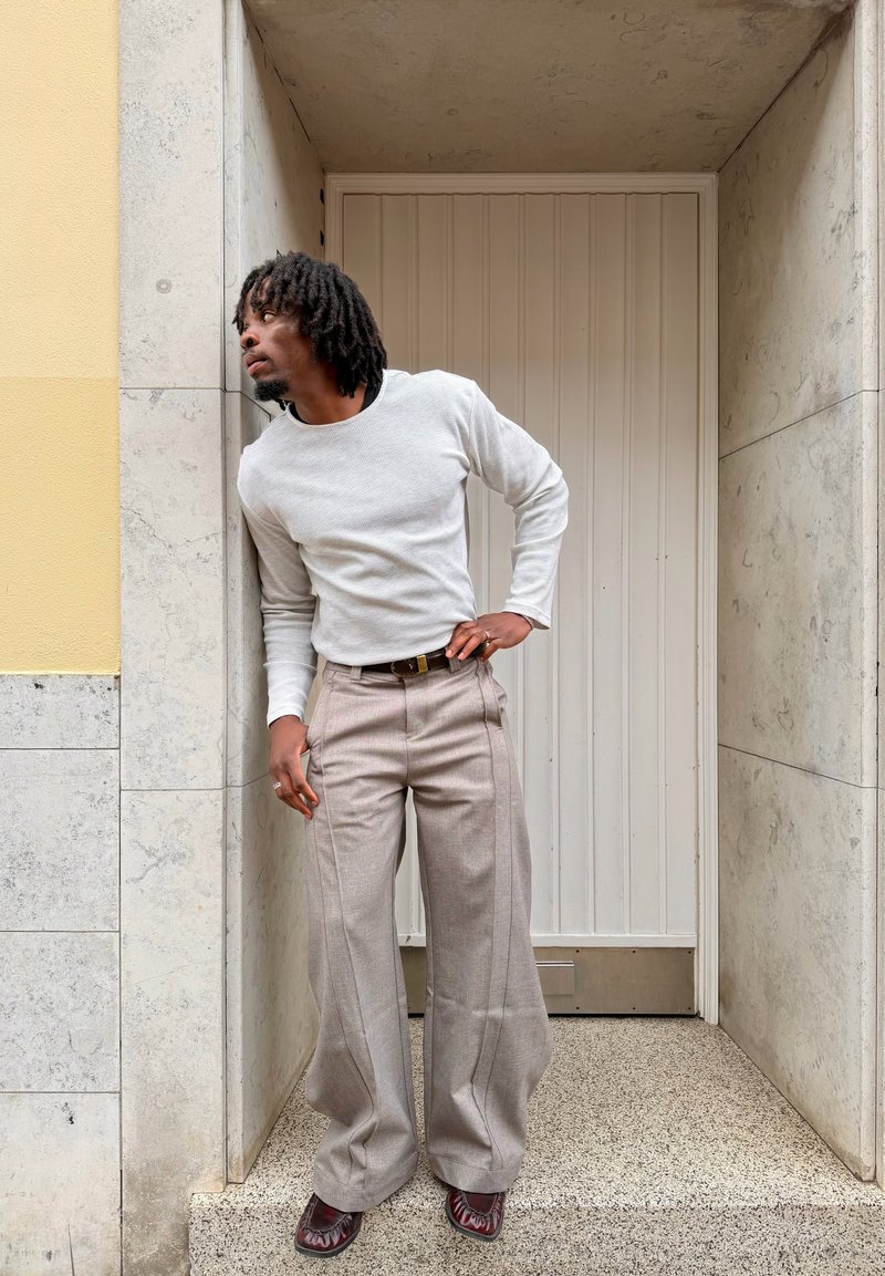 Homme avec des dreadlocks portant une chemise blanche à manches longues et un pantalon beige à jambes larges, appuyé contre une porte en pierre, regardant vers la gauche.