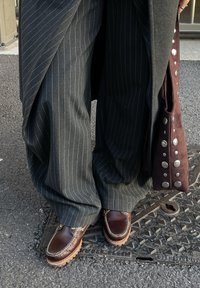 Jambes portant un pantalon large gris à rayures et des mocassins en cuir marron, debout sur une grille métallique, tenant un sac en daim marron avec des clous argentés.