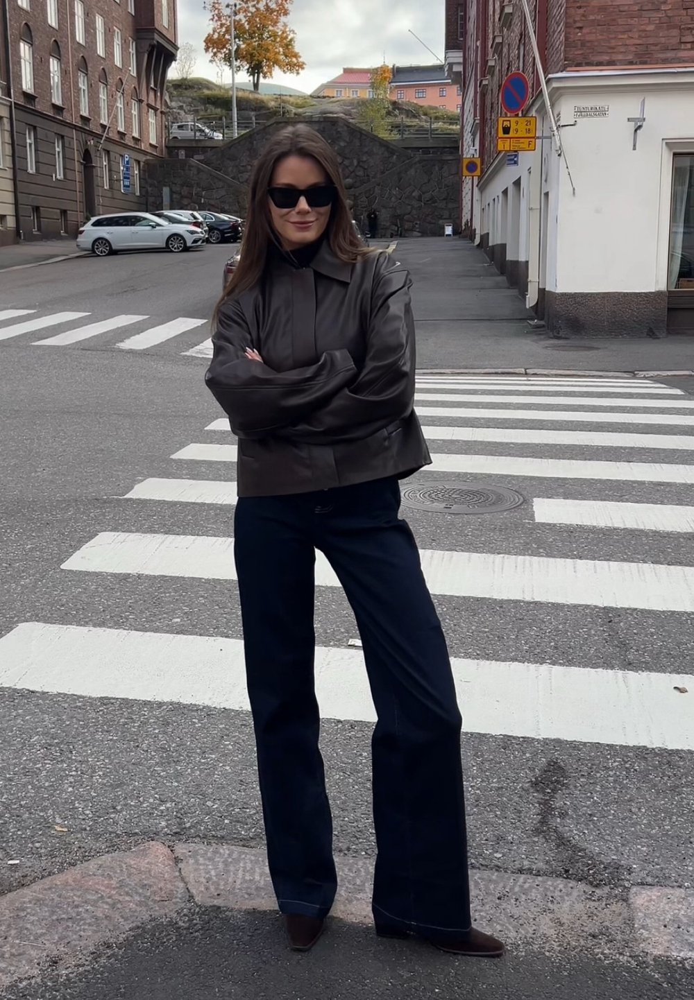 Jacket din piele neagră cu guler înalt, pantaloni largi de culoare albastră închisă și cizme cu toc maro, stând pe o trecere de zebra.