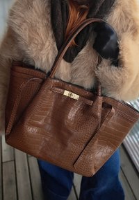 Personne portant un manteau en fourrure beige et des gants noirs, tenant un grand sac à main en cuir marron à motif crocodile avec une fermeture en or.