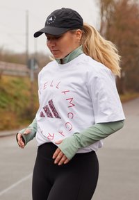 T-shirt blanc à manches courtes avec texte rouge et logo Adidas coloré, superposé sur un haut à manches longues vert, associé à des leggings noirs.