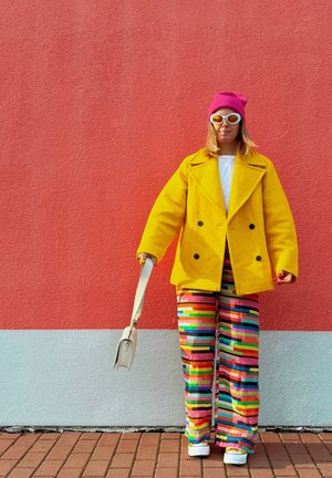 Abrigo amarillo cruzado de doble botonadura, top blanco, pantalones amplios de pierna ancha a rayas multicolores, gorro de punto rosa, gafas de sol blancas, llevando un bolso pequeño.