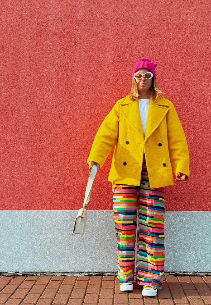 Manteau jaune à double boutonnage, haut blanc, pantalon large rayé multicolore, bonnet rose, lunettes de soleil blanches, portant un petit sac à main.