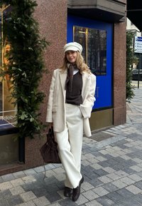 Manteau surdimensionné couleur crème, pull marron, pantalon blanc et chaussures marron foncé. Accessoirisé d'une écharpe et d'une casquette, tenant un sac foncé.