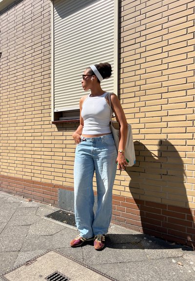 Camiseta blanca de tirantes, jeans anchos de un azul claro, zapatillas marrones y moradas, y un bolso tote beige. Las gafas de sol y la diadema blanca completan el look.