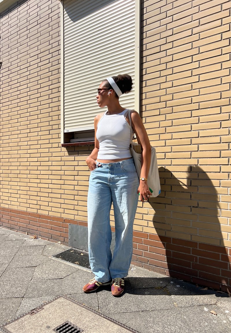 Canotta bianca, jeans a gamba larga azzurri chiari, sneakers marroni e viola, e una borsa tote beige. Gli occhiali da sole e la fascia bianca completano il look.