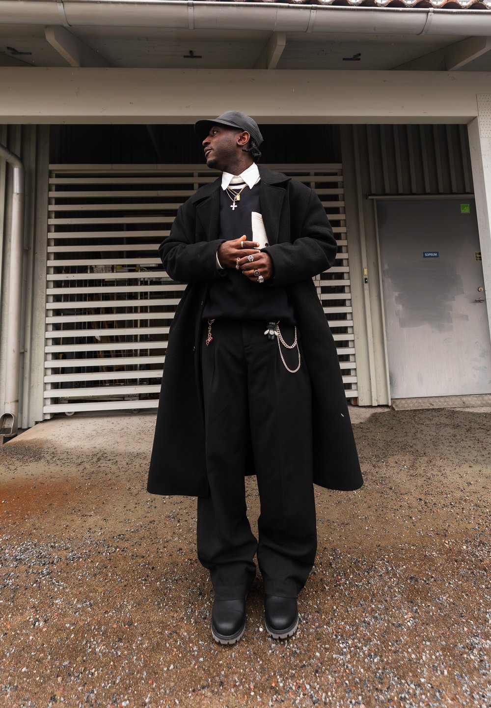 Homme portant un pardessus noir, un pantalon noir, des bottes et une casquette, se tenant les mains jointes devant un bâtiment avec une porte métallique et une grille d'aération.