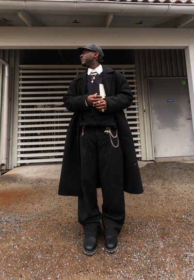 Hombre con abrigo negro, pantalones negros, botas y gorra, de pie con las manos entrelazadas, frente a un edificio con una puerta metálica y una rejilla de ventilación.