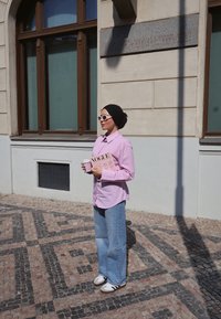 Rosa(stripete) skjorte, vide lyseblå jeans, hvite joggesko med svarte detaljer. Holder en avis og en kaffekopp, står på et mønstrete brosteinsgulv.
